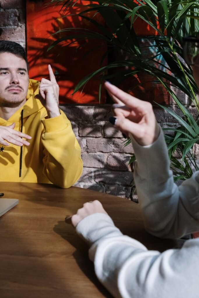 Two adults gesturing while sitting at a table indoors, emphasizing communication.